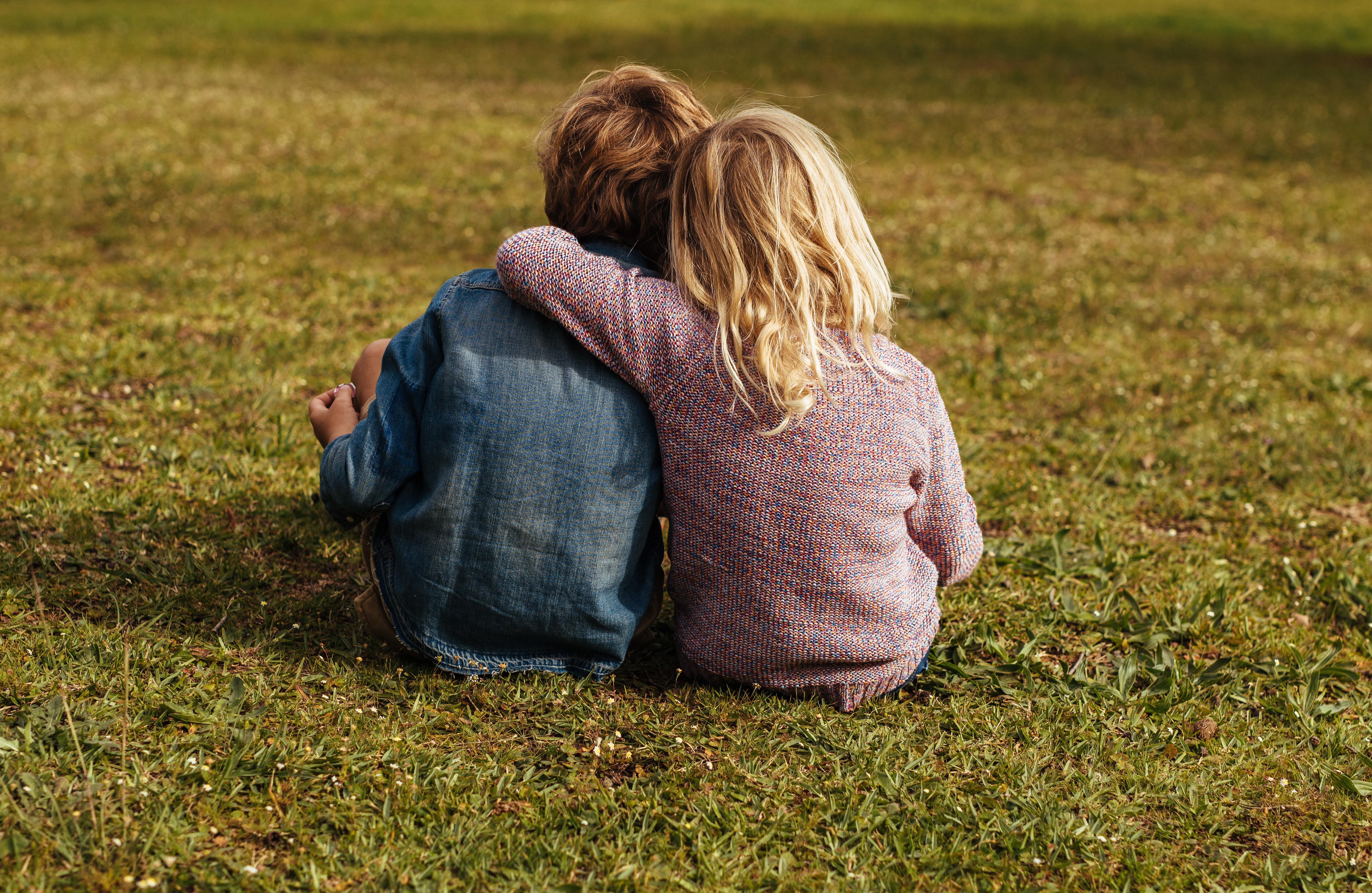 a brother and sister sitting in a field with their backs to the camera. The girl has her arm around the boy's shoulder