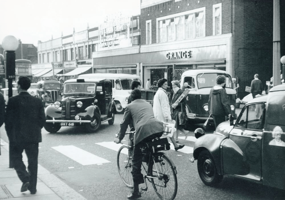 A black and white photo of a busy high street in the 1960s, with classic cars and pedestrians on a zebra crossing