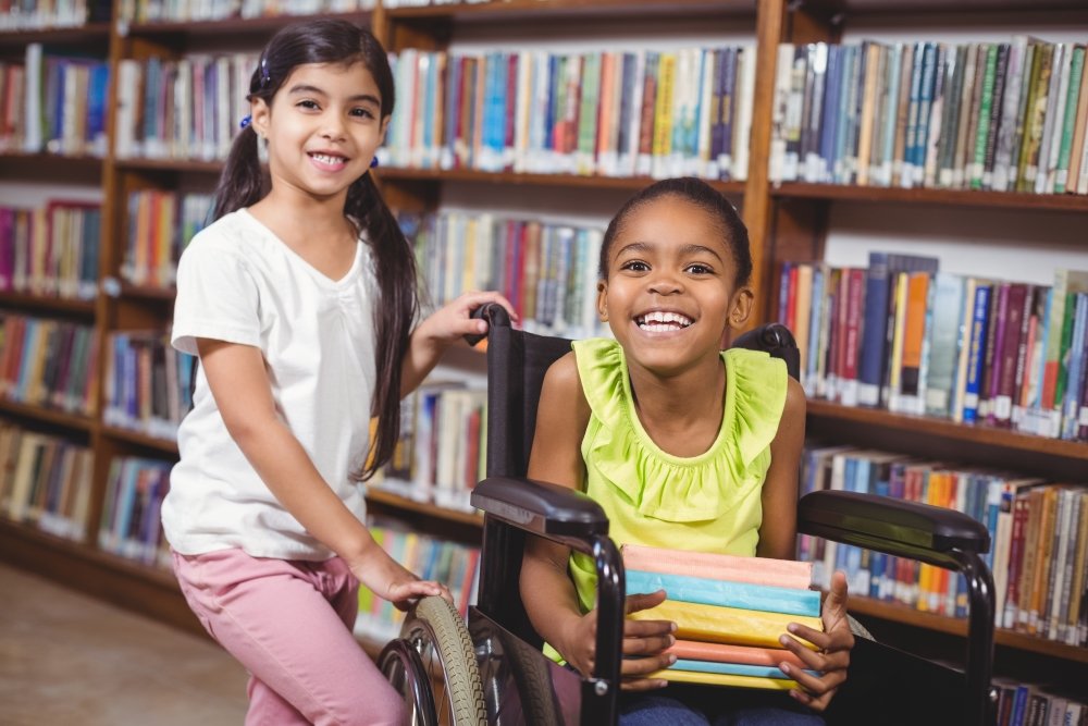 One child standing next to another in a wheelchair in a library